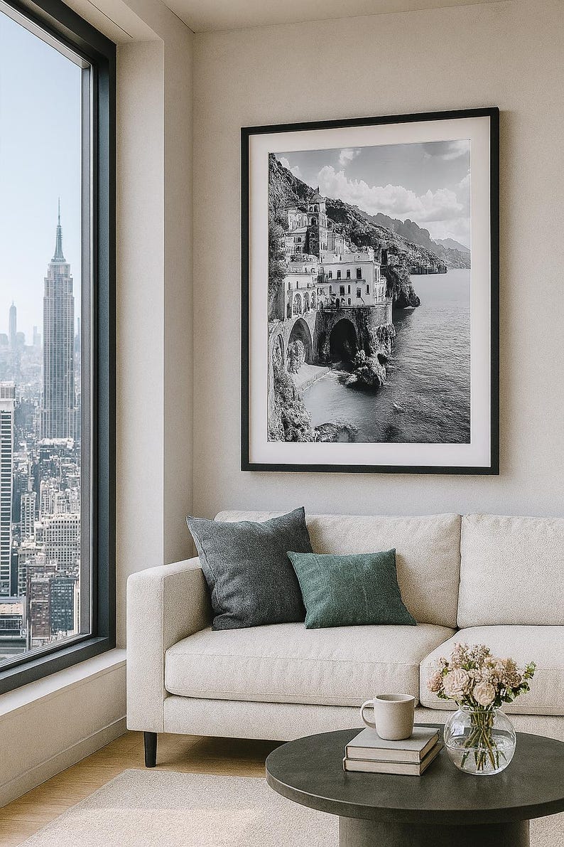 May include: A black and white framed print of a coastal town hangs above a beige sofa with two throw pillows. A round coffee table holds books, a mug, and a vase of flowers. A large window offers a city view.