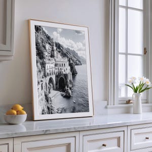 May include: A framed black and white photograph of a coastal Italian town. The image features a building on a cliffside, with arches and a tower, overlooking the sea. The frame is gold-colored, and the photograph is displayed on a white countertop.