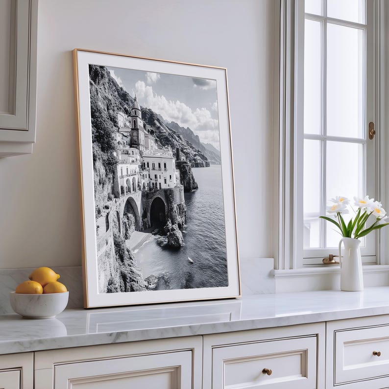 May include: A framed black and white photograph of a coastal Italian town. The image features a building on a cliffside, with arches and a tower, overlooking the sea. The frame is gold-colored, and the photograph is displayed on a white countertop.