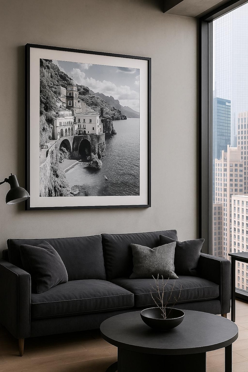 May include: A framed black and white photograph of a coastal town with buildings on a cliffside, a beach, and the sea. The photograph hangs above a dark gray sofa with throw pillows and a round coffee table. A window shows a city skyline.