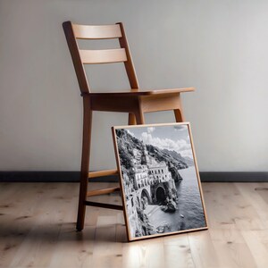 May include: A framed black and white photograph of a coastal Italian town, leaning against a wooden chair. The photograph features a building on a cliffside with arches and a beach. The chair is made of light-colored wood.