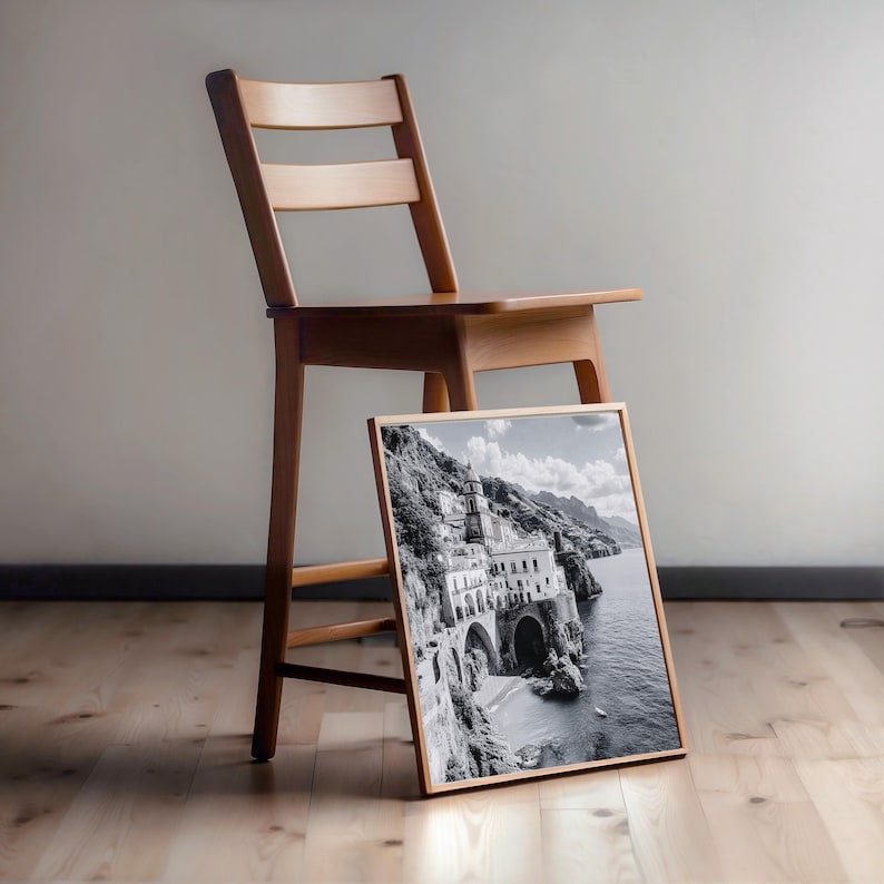 May include: A framed black and white photograph of a coastal Italian town, leaning against a wooden chair. The photograph features a building on a cliffside with arches and a beach. The chair is made of light-colored wood.