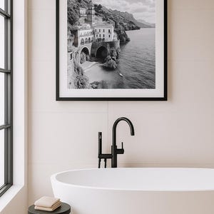 May include: Black and white framed print of a coastal Italian town. The image shows buildings on a cliffside, with arches and a beach. The print is displayed above a white bathtub with a black faucet and a small black side table.