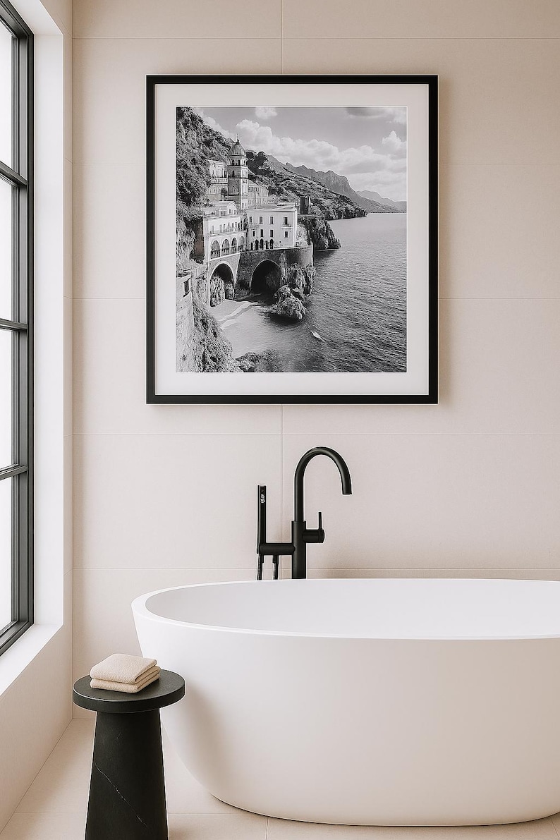 May include: Black and white framed print of a coastal Italian town. The image shows buildings on a cliffside, with arches and a beach. The print is displayed above a white bathtub with a black faucet and a small black side table.