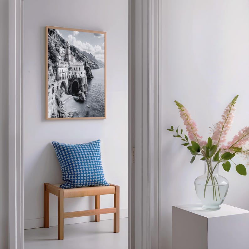 May include: A black and white framed print of a coastal Italian town hangs above a wooden bench with a blue patterned pillow. A clear glass vase with pink flowers sits on a white cube.