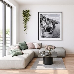 May include: A black and white framed print of a coastal town with arched structures and buildings on a cliffside. The print hangs above a modern gray sectional sofa with decorative pillows. A small black coffee table and rug complete the living room.