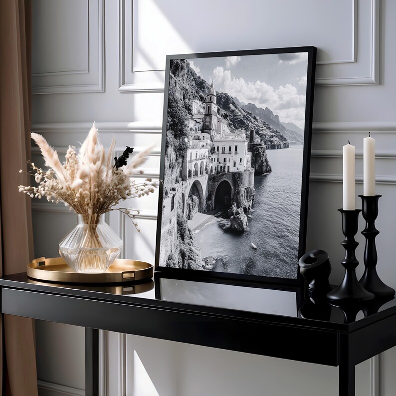 May include: Black and white framed print of a coastal town with buildings on a cliffside. The print is displayed on a black console table, alongside a vase of dried flowers, a gold tray, and black candle holders with white candles.