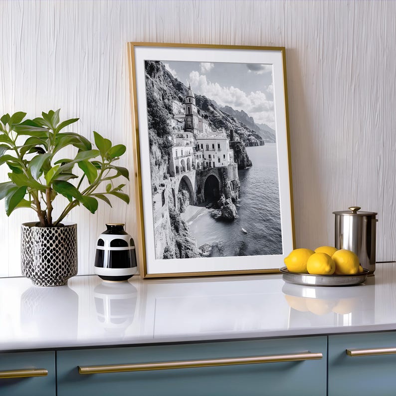 May include: A framed black and white print of a coastal Italian town with a gold frame. The image is displayed on a white countertop with a plant in a patterned pot, a black and white vase, and a plate of lemons.