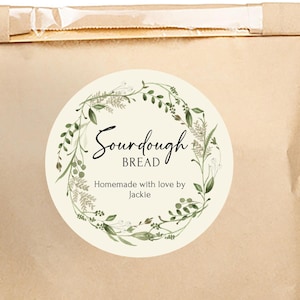 May include: A brown paper bag sealed with a clear tape strip. A round sticker on the front reads "Sourdough Bread" in script, surrounded by a green floral wreath. The text "Homemade with love by Jackie" is below.