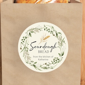 May include: A loaf of sourdough bread peeks out of a brown paper bag. The bag has a round sticker with the words "Sourdough Bread" and a floral wreath design. The bag rests on a wooden cutting board.