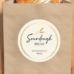 May include: A brown paper bag with a round sticker that says "Sourdough Bread from the kitchen of Maria". The sticker has a gold circle around it with wheat stalks inside. A loaf of sourdough bread is sitting on top of the bag.