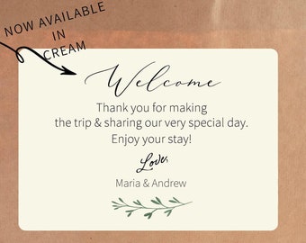 May include: A cream-colored welcome sign with a handwritten font that says "Welcome". The sign also includes the text "Thank you for making the trip & sharing our very special day. Enjoy your stay! Love, Maria & Andrew". The sign is decorated with a small green sprig.