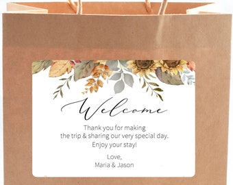 May include: A brown paper gift bag with a white rectangular label. The label features a floral design with sunflowers and autumn leaves. The word "Welcome" is written in elegant script, with a message below.