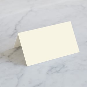 May include: A folded, off-white place card, perfect for weddings or events. The card is rectangular with a simple, elegant design. The card is blank, ready for personalization. The background is a marble surface.