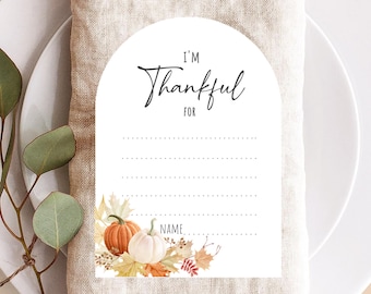 May include: White place card with an arched top, featuring the text "I'M Thankful FOR" in black script. The card includes lines for writing and is decorated with watercolor pumpkins and fall leaves. A beige napkin and white plate are in the background.