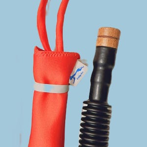 May include: A red wine bottle bag with two handles and a black wine aerator with a wooden top. The bag has a white band and a small tag with the text "Tally-Go".