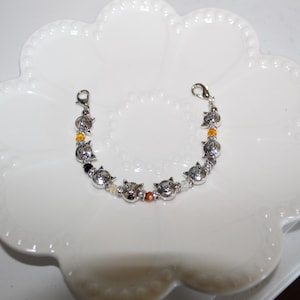 May include: A silver-toned bracelet featuring cat-shaped charms and colorful beads. The bracelet is displayed on a white, scalloped-edge plate. The charms are detailed with facial features, and the beads are orange, black, and clear.