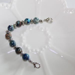 May include: A beaded bracelet featuring round, multi-colored stone beads in shades of blue, teal, and brown, interspersed with small, ornate silver-toned metal spacers. The bracelet is finished with a silver-toned lobster claw clasp at each end.