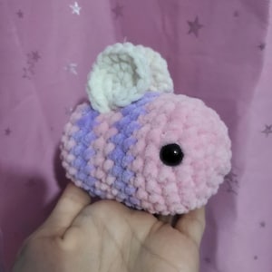 May include: A handmade, plush bee toy in shades of pink and lavender, with a white head and black eyes. The bee is made of soft, textured yarn, and is held in a hand.