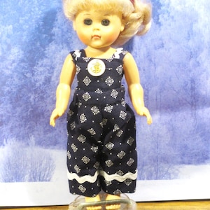 May include: A blonde doll with a red headband wearing dark blue overalls featuring a white floral pattern and a white wavy trim at the hem. The overalls have a single white button on the chest. The doll stands on a clear plastic heart-shaped base.