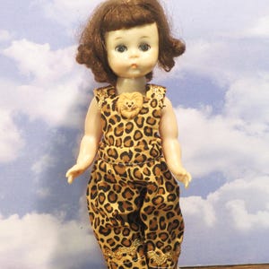 May include: A doll with brown hair and blue eyes wearing a sleeveless leopard print jumpsuit. The jumpsuit features a small tan lion-shaped embellishment at the neckline and gold-colored trim along the bottom of the pant legs. The doll stands on a clear base.