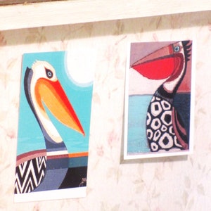 K&ouml;nnte beinhalten: Zwei bunte Vogel-Illustrationen auf wei&szlig;em Karton. Die linke Illustration ist ein Pelikan mit einem blauen, orangefarbenen und schwarzen Muster. Die rechte Illustration ist ein Vogel mit einem roten Schnabel und einem schwarz-wei&szlig;en Muster.