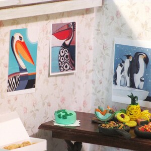K&ouml;nnte beinhalten: Eine Miniatur-Puppenhausszene mit einem Tisch, der f&uuml;r eine Party gedeckt ist. Es gibt mehrere kleine Schalen mit Essen, darunter eine Ananas, eine gr&uuml;ne Torte und eine Schale mit Garnelen. Die W&auml;nde sind mit gerahmten Bildern von V&ouml;geln dekoriert.