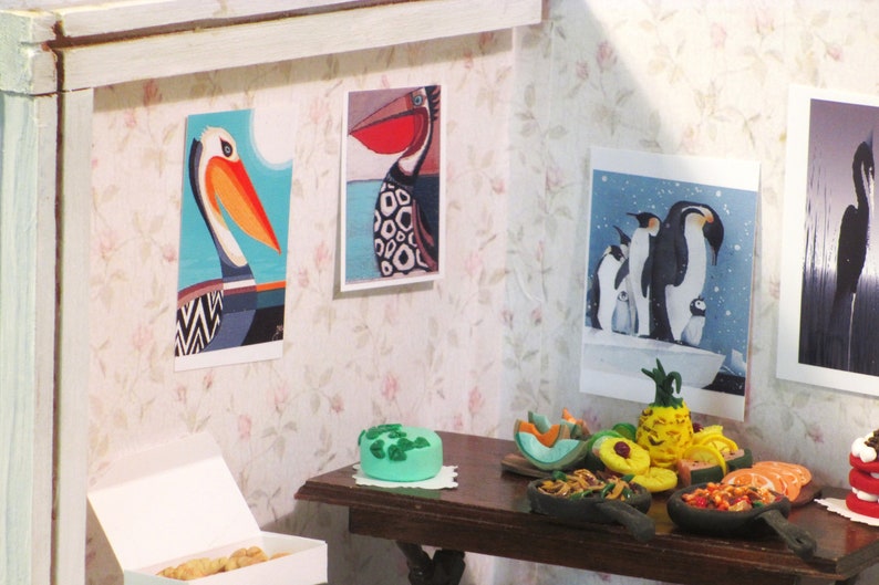 K&ouml;nnte beinhalten: Eine Miniatur-Puppenhausszene mit einem Tisch, der f&uuml;r eine Party gedeckt ist. Es gibt mehrere kleine Schalen mit Essen, darunter eine Ananas, eine gr&uuml;ne Torte und eine Schale mit Garnelen. Die W&auml;nde sind mit gerahmten Bildern von V&ouml;geln dekoriert.
