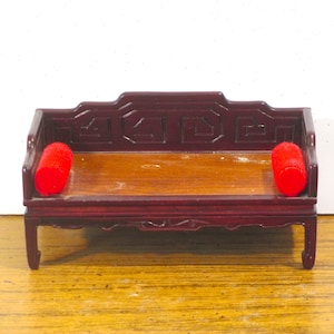 May include: Miniature wooden bench with a dark red finish and a light brown seat. The bench has a decorative back and two red cylindrical pillows. A small piece of furniture, likely for a dollhouse or display.