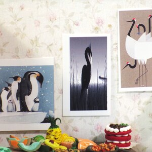 K&ouml;nnte beinhalten: Drei gerahmte Drucke von Tierillustrationen. Der erste Druck zeigt eine Gruppe von Pinguinen vor einem schneebedeckten Hintergrund. Der zweite Druck zeigt einen schwarzen Vogel, der in einem Feld mit hohem Gras steht. Der dritte Druck zeigt eine Gruppe von wei&szlig;en Kranichen mit roten K&ouml;pfen, die auf einem braunen Hintergrund stehen.