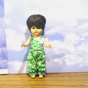 May include: A small doll with short black hair wearing a green jumpsuit featuring a white floral pattern. The jumpsuit has a white flower accent on the left shoulder and a dark green zigzag trim along the bottom hem of the pant legs.