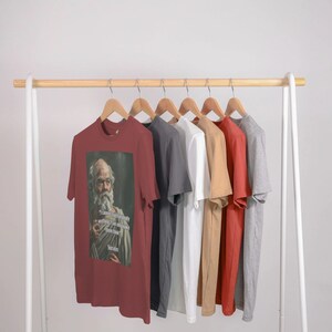 May include: A rack of t-shirts in various colors, including red, white, gray, beige, and orange. The red t-shirt features a printed image of a man with a long white beard and the text "I learned everything I needed to know in kindergarten."