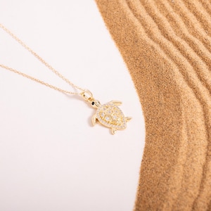 Puede incluir: Un collar dorado con un colgante de tortuga. El colgante está adornado con pequeñas piedras preciosas transparentes y un ojo negro. El collar se muestra sobre una superficie blanca con una textura arenosa en el fondo.