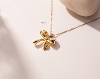 Puede incluir: Un collar con colgante de lazo dorado con pequeños detalles de perlas. El lazo es el punto focal, suspendido de una delicada cadena. El collar se exhibe sobre una superficie blanca con una piedra en el fondo.