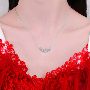 May include: A silver necklace with a row of small, clear cubic zirconia stones set in a curved line. The necklace is on a model wearing a red dress.