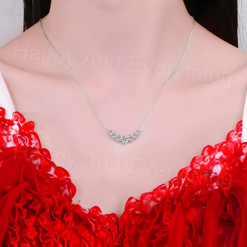 May include: A silver necklace with a row of small, clear cubic zirconia stones set in a curved line. The necklace is on a model wearing a red dress.