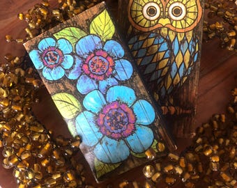 May include: Two vintage wooden boxes with painted designs. One box features blue flowers with purple centers and green leaves. The other box has a colorful owl design. A beaded garland surrounds the boxes.