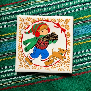 May include: A white ceramic tile with a festive Christmas scene. A young boy in a red and green hat and blue pants carries a bundle of logs. A small brown dog walks beside him. The tile is decorated with a red and gold wreath and green leaves.