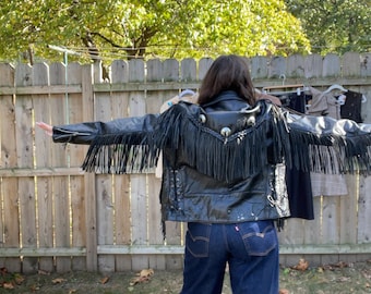 May include: Black leather jacket with fringe detailing and silver studs. The jacket has a classic biker style with long fringe along the back and sleeves. The person is wearing dark blue denim jeans.