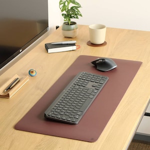 Könnte beinhalten: Eine braune Leder-Schreibtischunterlage mit einer schwarzen Tastatur und Maus. Die Unterlage liegt auf einem Holztisch mit einer Pflanze, einer Tasse und einem Stifthalter.