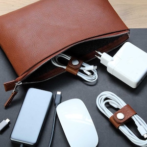 Könnte beinhalten: Eine braune Leder-Reißverschlusstasche mit schwarzem Futter. Die Tasche ist offen und enthält ein weißes Netzteil, eine weiße Maus und mehrere weiße Kabel mit braunen Leder-Kabelorganisatoren.
