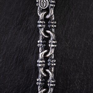 May include: Close-up of a silver chain necklace with a distinctive bamboo-style design. The chain links are intricately detailed, and the top features a circular emblem. The necklace is displayed against a dark, textured background.