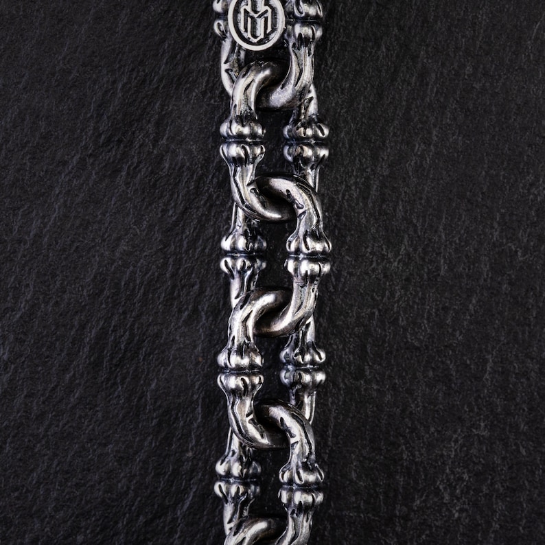 May include: Close-up of a silver chain necklace with a distinctive bamboo-style design. The chain links are intricately detailed, and the top features a circular emblem. The necklace is displayed against a dark, textured background.