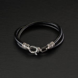 Könnte beinhalten: Ein schwarzes Lederarmband mit einem silbernen Verschluss. Der Verschluss ist mit filigranen Details verziert und mit kleinen silbernen Perlen besetzt.