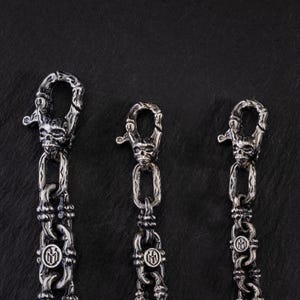 May include: Three silver metal chains with skull-shaped clasps. The chains have a detailed link design and a circular emblem. The background is a dark, textured surface.