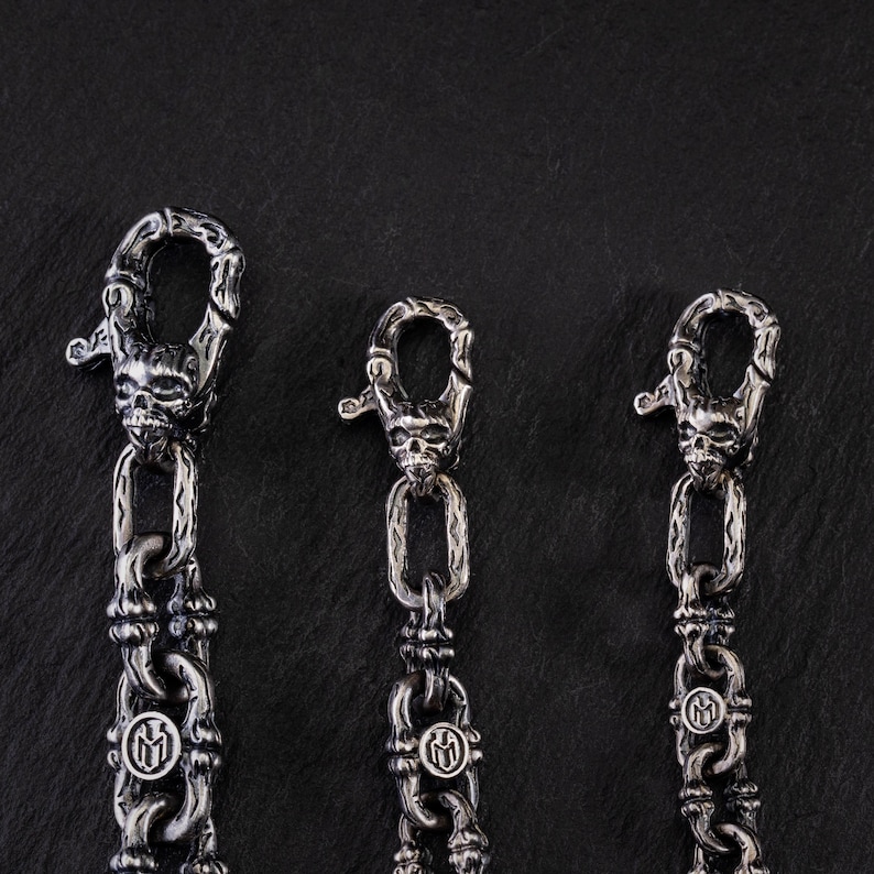 May include: Three silver metal chains with skull-shaped clasps. The chains have a detailed link design and a circular emblem. The background is a dark, textured surface.