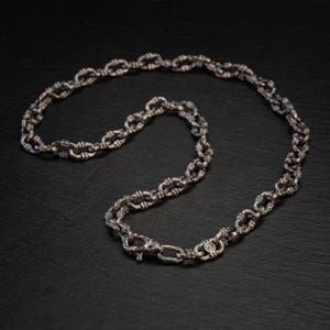May include: A silver necklace featuring an ornate chain pattern. The chain links are oval and joined by decorative knots. The necklace is presented on a dark surface, accentuating its metallic shine. The clasp is visible.