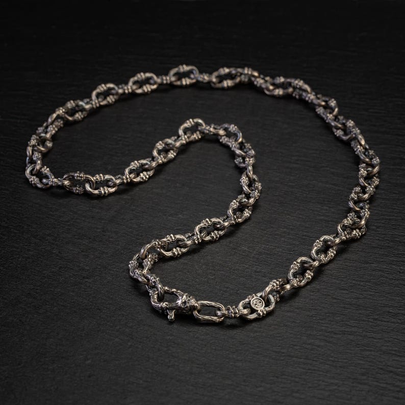 May include: A silver necklace featuring an ornate chain pattern. The chain links are oval and joined by decorative knots. The necklace is presented on a dark surface, accentuating its metallic shine. The clasp is visible.