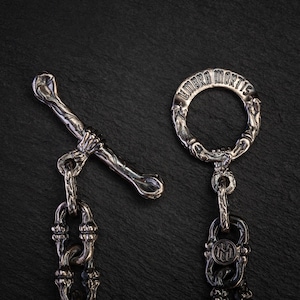 May include: Close-up of a silver metal clasp and chain. The clasp has a bone-shaped bar held by a hand, and a ring with the inscription "UMBRA MORTIS". The chain has detailed links. The background is dark.