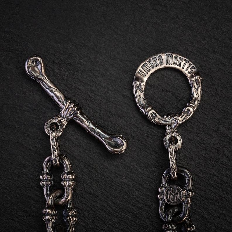 May include: Close-up of a silver metal clasp and chain. The clasp has a bone-shaped bar held by a hand, and a ring with the inscription "UMBRA MORTIS". The chain has detailed links. The background is dark.